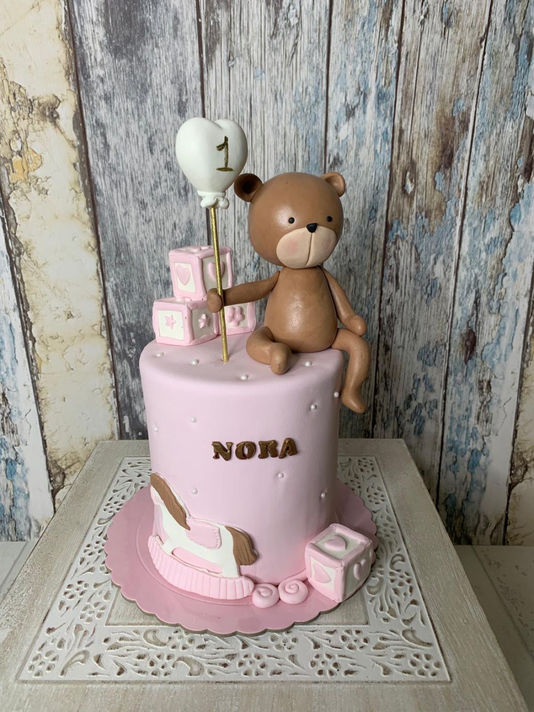 Tarta fondant para primer cumpleaños de niña con figura de oso, globo de corazón y bloques de juguete.