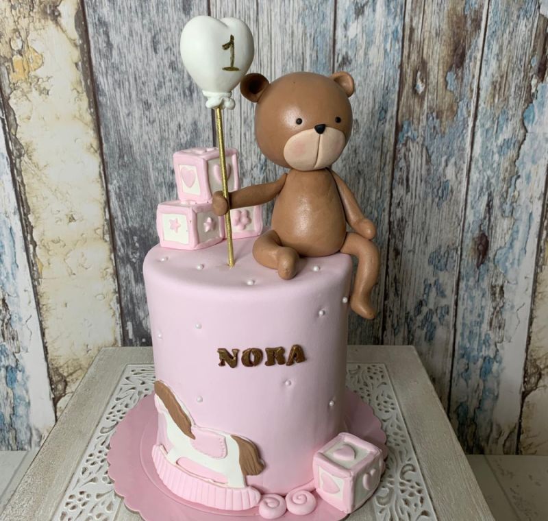 Tarta fondant para primer cumpleaños de niña con figura de oso, globo de corazón y bloques de juguete.